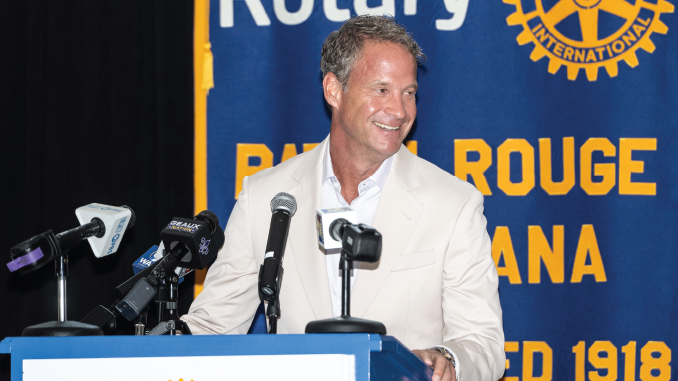 Man in a light beige suit smiles at a podium with multiple microphones, standing in front of a blue and yellow Rotary banner.