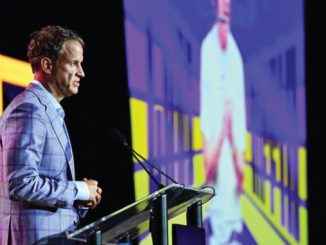Man in a light-blue plaid suit speaks at a podium on a stage with a large screen behind him.