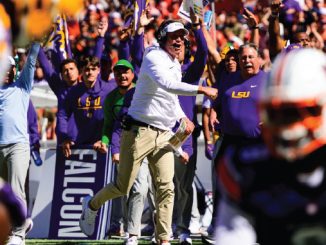 LSU coach in white shirt and headset celebrating on the sideline as purple-clad fans cheer wildly behind him.