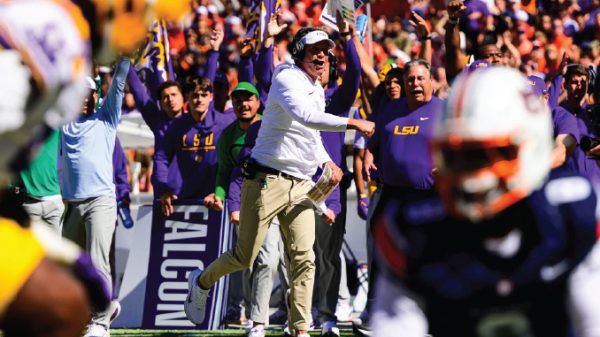 LSU coach in white shirt and headset celebrating on the sideline as purple-clad fans cheer wildly behind him.