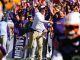 LSU coach in white shirt and headset celebrating on the sideline as purple-clad fans cheer wildly behind him.