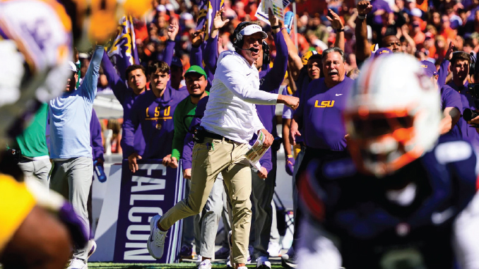 LSU coach in white shirt and headset celebrating on the sideline as purple-clad fans cheer wildly behind him.