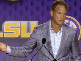 Man in a plaid gray suit at a podium, gesturing with his left hand, LSU backdrop behind him.
