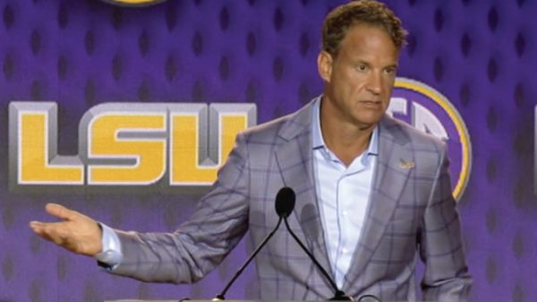 Man in a plaid gray suit at a podium, gesturing with his left hand, LSU backdrop behind him.