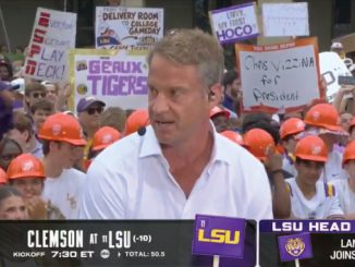 A man in a light blue shirt speaking into a headset mic in front of a crowded fan group with orange hard hats and handmade signs, on a college football broadcast overlay.