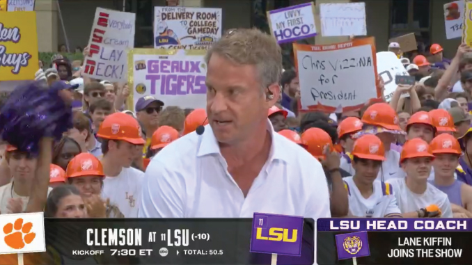 A man in a light blue shirt speaking into a headset mic in front of a crowded fan group with orange hard hats and handmade signs, on a college football broadcast overlay.