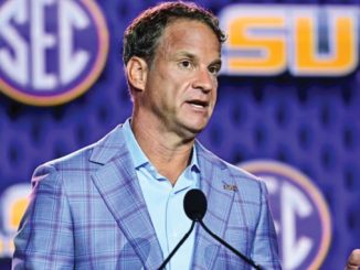 Man in a light blue shirt and blue plaid blazer speaks at a podium, with SEC and LSU logos in the background.