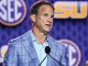 Man in a light blue shirt and blue plaid blazer speaks at a podium, with SEC and LSU logos in the background.