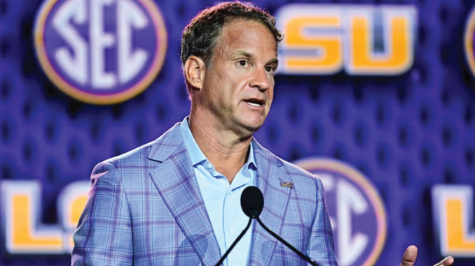Man in a light blue shirt and blue plaid blazer speaks at a podium, with SEC and LSU logos in the background.