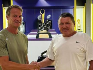 Two men shake hands in a trophy room with glass display cases and sports trophies in the background.