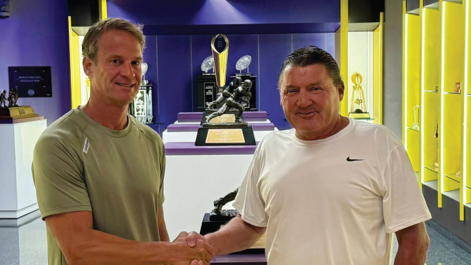 Two men shake hands in a trophy room with glass display cases and sports trophies in the background.