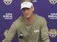 LSU coach in an olive hoodie and white LSU visor speaks at a press conference, microphone in front, backdrop with tiger logos and Farm Bureau branding.