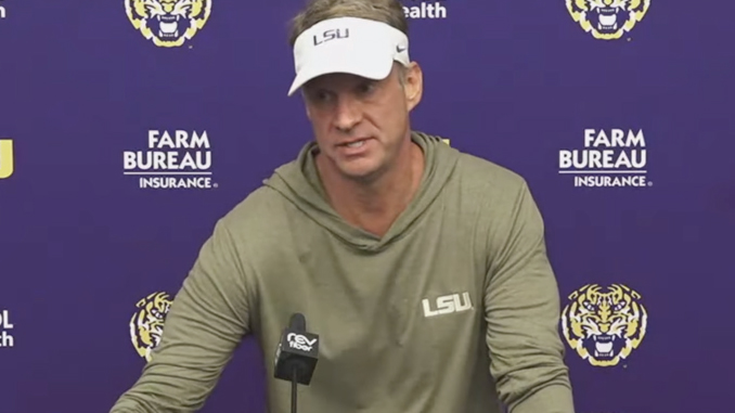 LSU coach in an olive hoodie and white LSU visor speaks at a press conference, microphone in front, backdrop with tiger logos and Farm Bureau branding.
