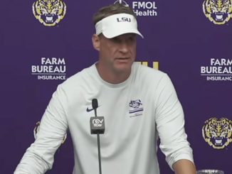 Man wearing a white LSU visor and shirt at a press conference, speaking into a microphone against a purple backdrop with LSU and Farm Bureau logos.