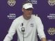 Man wearing a white LSU visor and shirt at a press conference, speaking into a microphone against a purple backdrop with LSU and Farm Bureau logos.