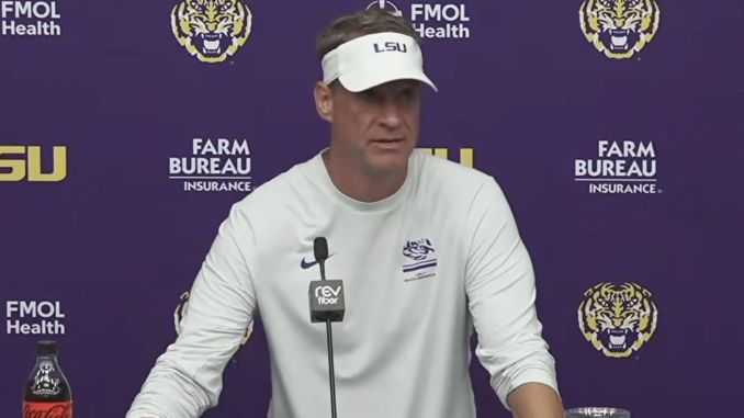 Man wearing a white LSU visor and shirt at a press conference, speaking into a microphone against a purple backdrop with LSU and Farm Bureau logos.