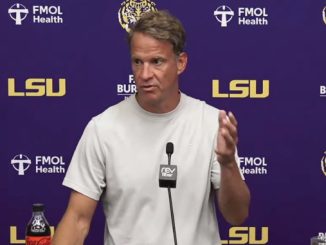 Male football coach giving a press conference at LSU, standing behind a microphone behind a podium.
