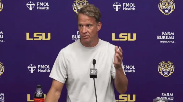 Male football coach giving a press conference at LSU, standing behind a microphone behind a podium.