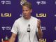 Male football coach giving a press conference at LSU, standing behind a microphone behind a podium.
