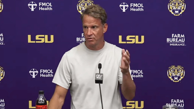 Male football coach giving a press conference at LSU, standing behind a microphone behind a podium.