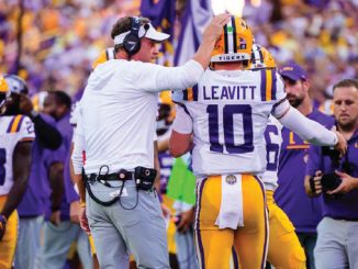 LSU coach adjusts quarterback #10 Leavitt's helmet on the sideline during a game.