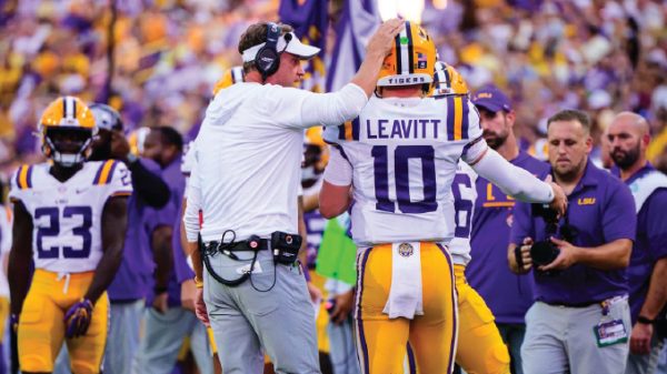 LSU coach adjusts quarterback #10 Leavitt's helmet on the sideline during a game.