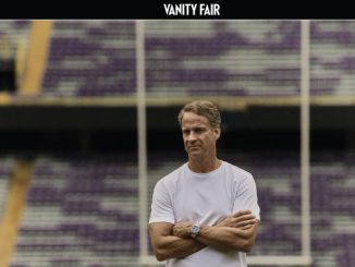 Man in a white T-shirt with arms crossed standing in an empty football stadium.