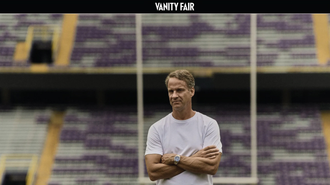 Man in a white T-shirt with arms crossed standing in an empty football stadium.