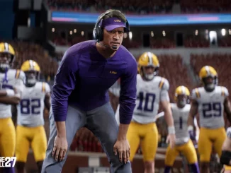 LSU football coach in a purple shirt and headset crouches on the field, surrounded by players in white and yellow uniforms during a game.
