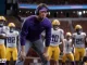 LSU football coach in a purple shirt and headset crouches on the field, surrounded by players in white and yellow uniforms during a game.