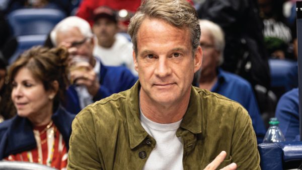 Man in an olive jacket sits at a panel, looking at the camera, with a blurred conference audience behind him.
