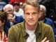 Man in an olive jacket sits at a panel, looking at the camera, with a blurred conference audience behind him.