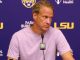 Man wearing a light purple shirt speaks into a microphone at a press conference with an LSU backdrop behind him.