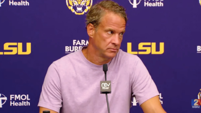 Man wearing a light purple shirt speaks into a microphone at a press conference with an LSU backdrop behind him.