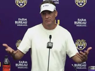 LSU coach in a white shirt speaking at a purple press conference backdrop with LSU and Farm Bureau logos, microphones in front of him