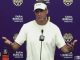 LSU coach in a white shirt speaking at a purple press conference backdrop with LSU and Farm Bureau logos, microphones in front of him