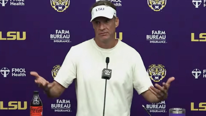 LSU coach in a white shirt speaking at a purple press conference backdrop with LSU and Farm Bureau logos, microphones in front of him