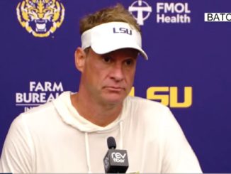 Man in a white hoodie and LSU visor sits at a press conference microphone in front of a blue backdrop with LSU logos.