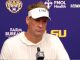 Man in a white hoodie and LSU visor sits at a press conference microphone in front of a blue backdrop with LSU logos.