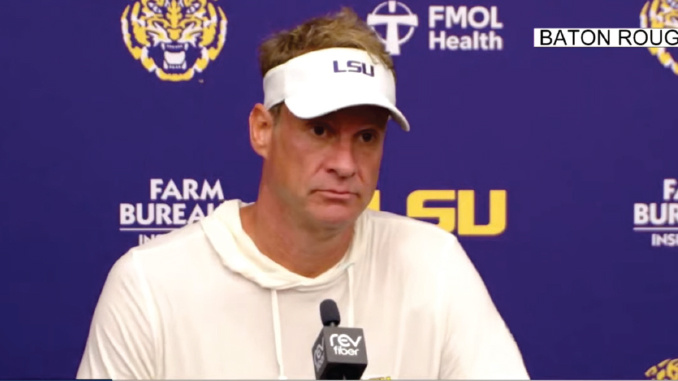 Man in a white hoodie and LSU visor sits at a press conference microphone in front of a blue backdrop with LSU logos.