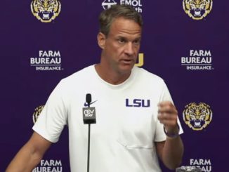LSU football coach at a press conference, wearing a white shirt with LSU, speaking into a microphone against a purple backdrop with LSU logos.