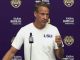 LSU football coach at a press conference, wearing a white shirt with LSU, speaking into a microphone against a purple backdrop with LSU logos.