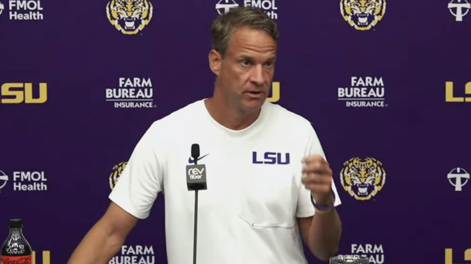 LSU football coach at a press conference, wearing a white shirt with LSU, speaking into a microphone against a purple backdrop with LSU logos.