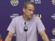 Coach at a press conference behind a podium with a microphone, wearing a light purple shirt, purple backdrop with LSU logos.