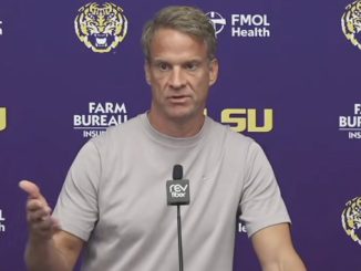 Man in a light gray shirt speaks at a press conference, gesturing with his left hand, against a purple backdrop with LSU logos and Farm Bureau branding.