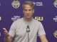 Man in a light gray shirt speaks at a press conference, gesturing with his left hand, against a purple backdrop with LSU logos and Farm Bureau branding.