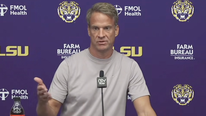 Man in a light gray shirt speaks at a press conference, gesturing with his left hand, against a purple backdrop with LSU logos and Farm Bureau branding.