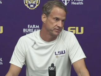 LSU coach speaks at a press conference, wearing a white LSU shirt, seated behind a microphone, with a purple backdrop featuring LSU logos.