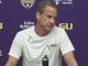 LSU coach speaks at a press conference, wearing a white LSU shirt, seated behind a microphone, with a purple backdrop featuring LSU logos.