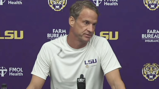 LSU coach speaks at a press conference, wearing a white LSU shirt, seated behind a microphone, with a purple backdrop featuring LSU logos.